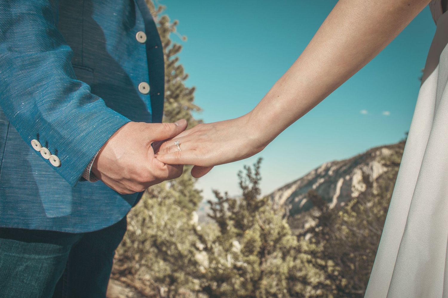 Couple holding hands outdoors during wedding ceremony with mountain and blue sky backdrop.