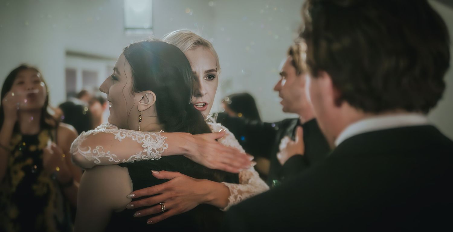 Two women share an emotional hug at a wedding reception, with guests and soft bokeh lights in the background.
