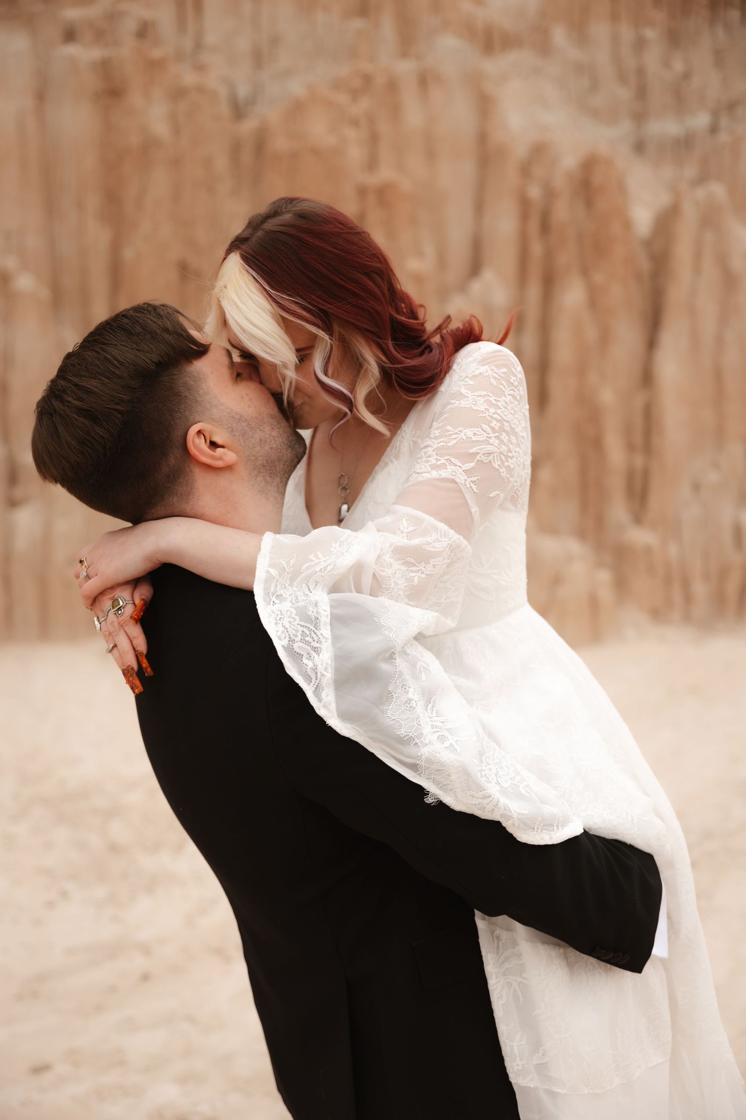 Groom lifts and kisses bride in white lace wedding gown against sandy desert rock formation backdrop.