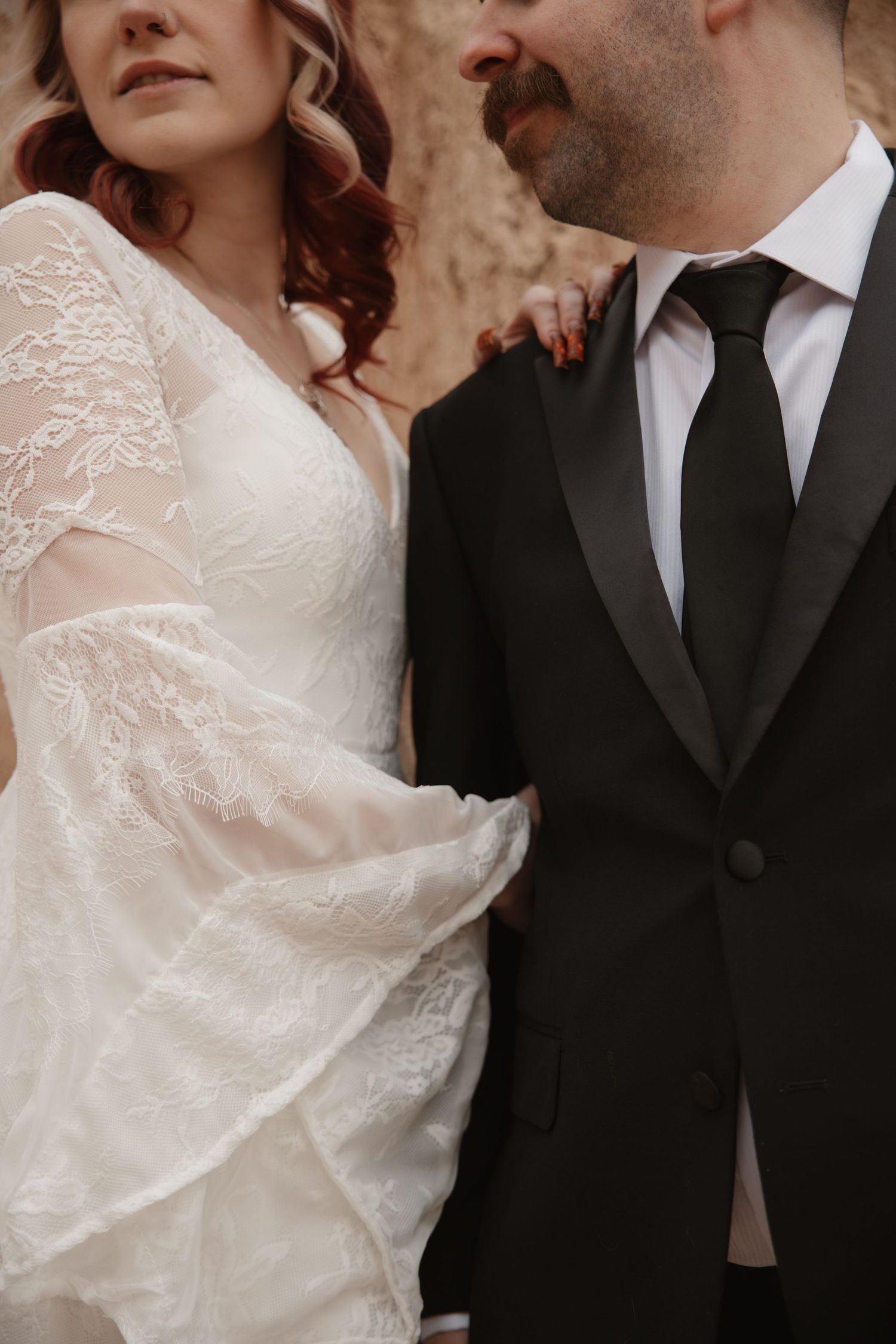 Bride in lace wedding dress stands beside groom in black tuxedo during outdoor wedding photo session.