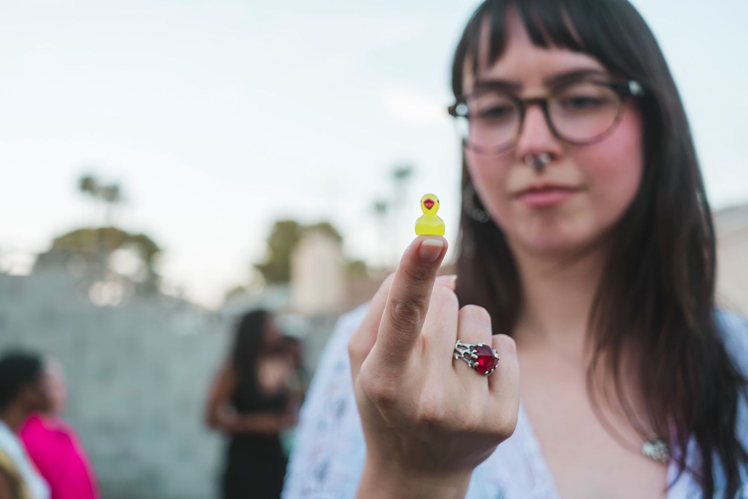 Woman holding up finger with tiny yellow rubber duck figurine outdoors at a social gathering.