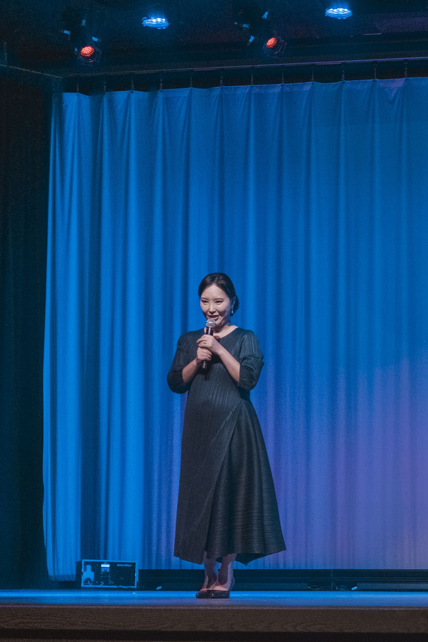 Woman in dark dress holds microphone on blue-lit stage with curtain backdrop.