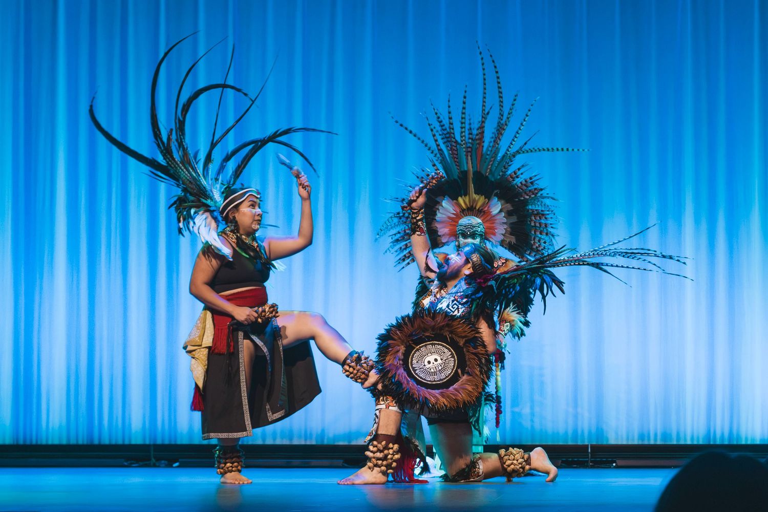 Two performers in elaborate Aztec feathered headdresses and traditional costumes dance on a blue-lit stage.