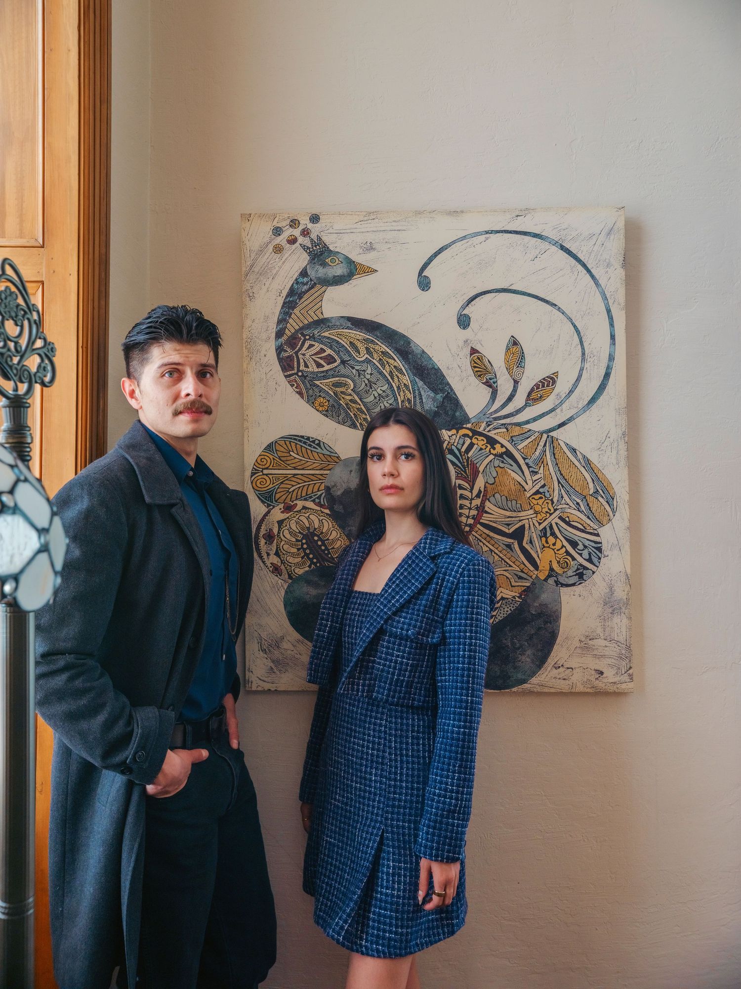 A man and woman in blue outfits stand before a decorative peacock painting in an elegant interior.