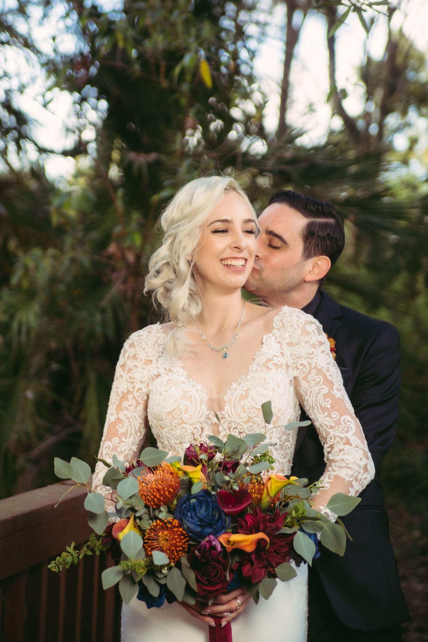 Groom kisses smiling bride on cheek as she holds colorful bouquet in lace long-sleeve wedding gown outdoors.