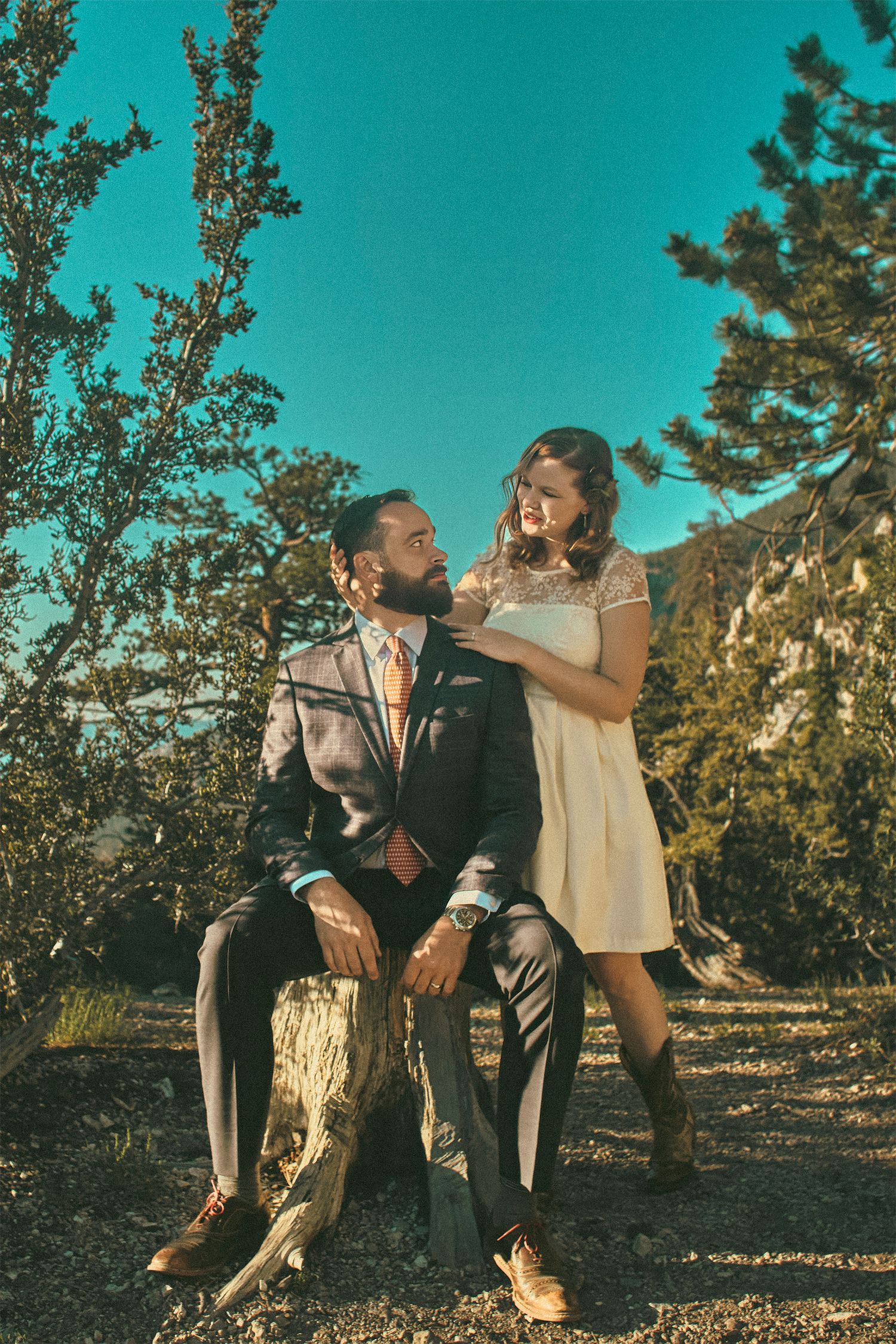 Couple in wedding attire posing outdoors on a tree stump surrounded by pine trees and rocky mountain landscape.