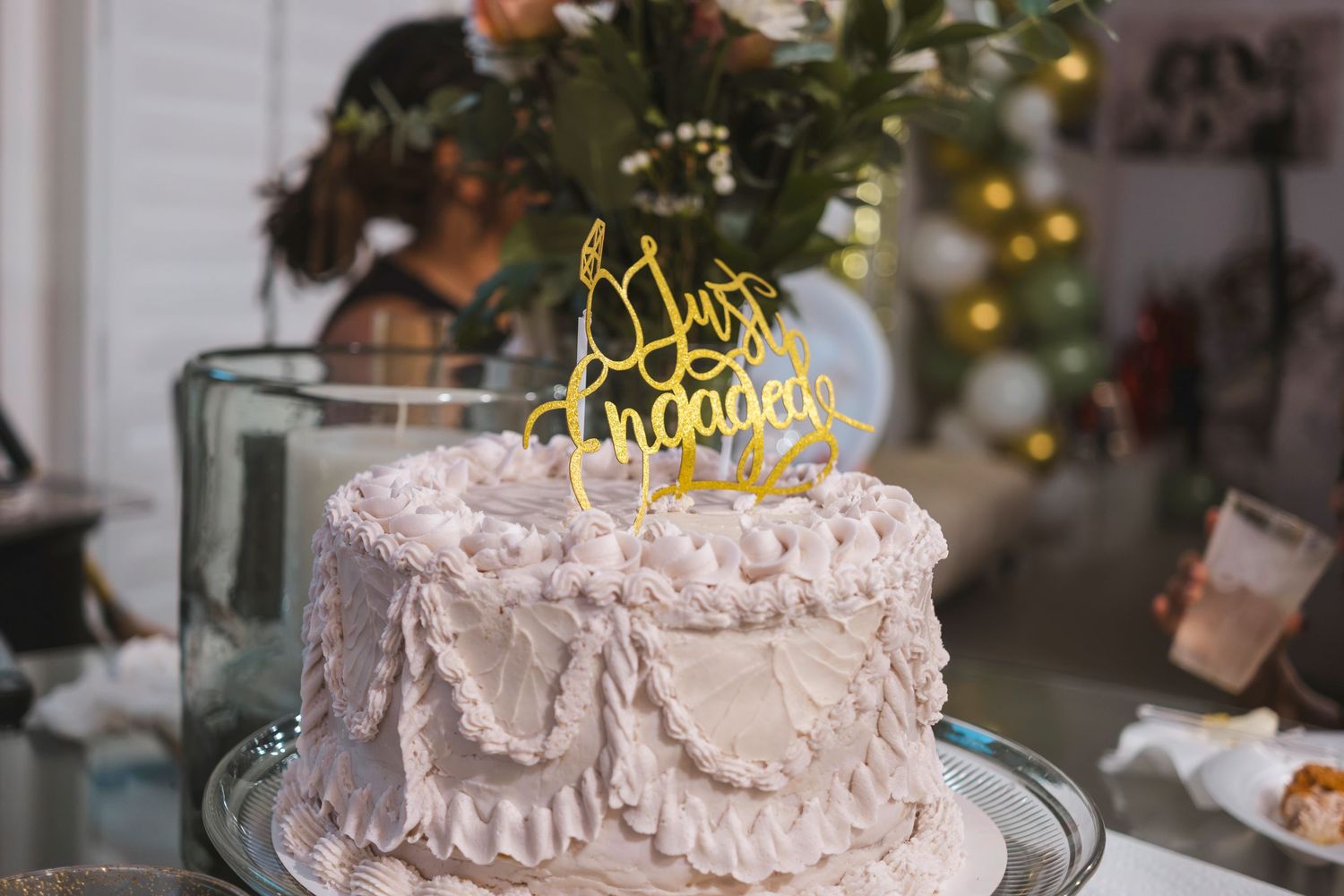 White engagement cake with gold 'Just Engaged' topper on glass stand, decorated with ruffled frosting.