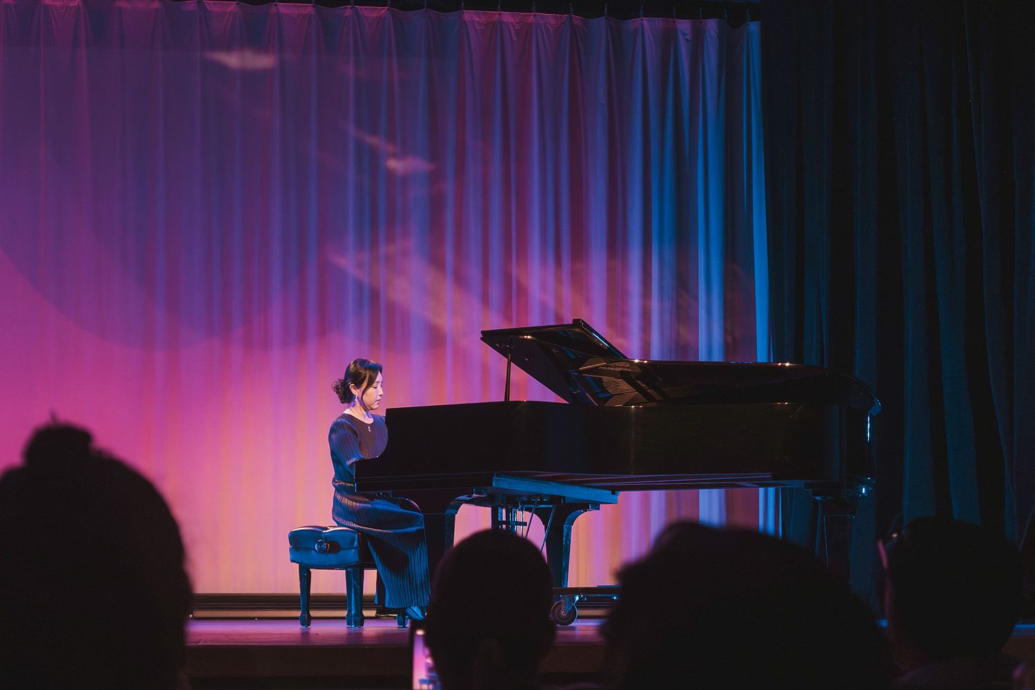 A pianist performs solo on a grand piano on a dramatic stage lit with pink and blue lighting.