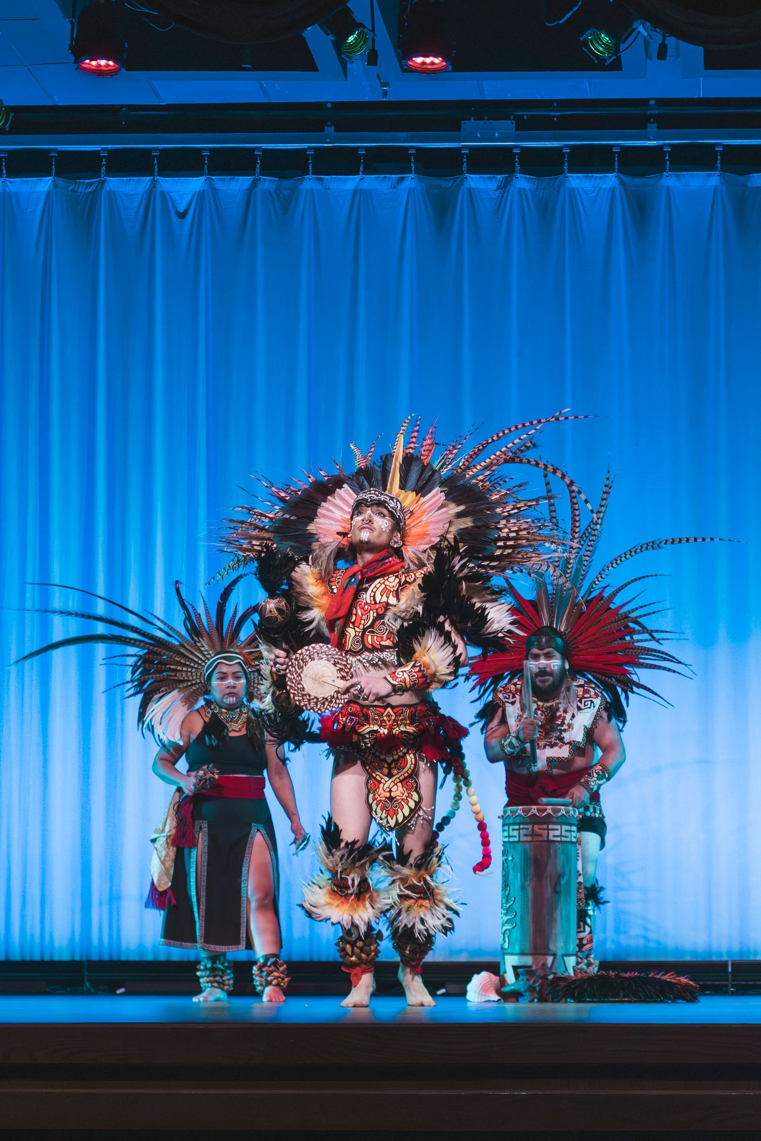 Three performers in elaborate Aztec feathered headdresses and traditional costumes dance on a blue-lit stage.