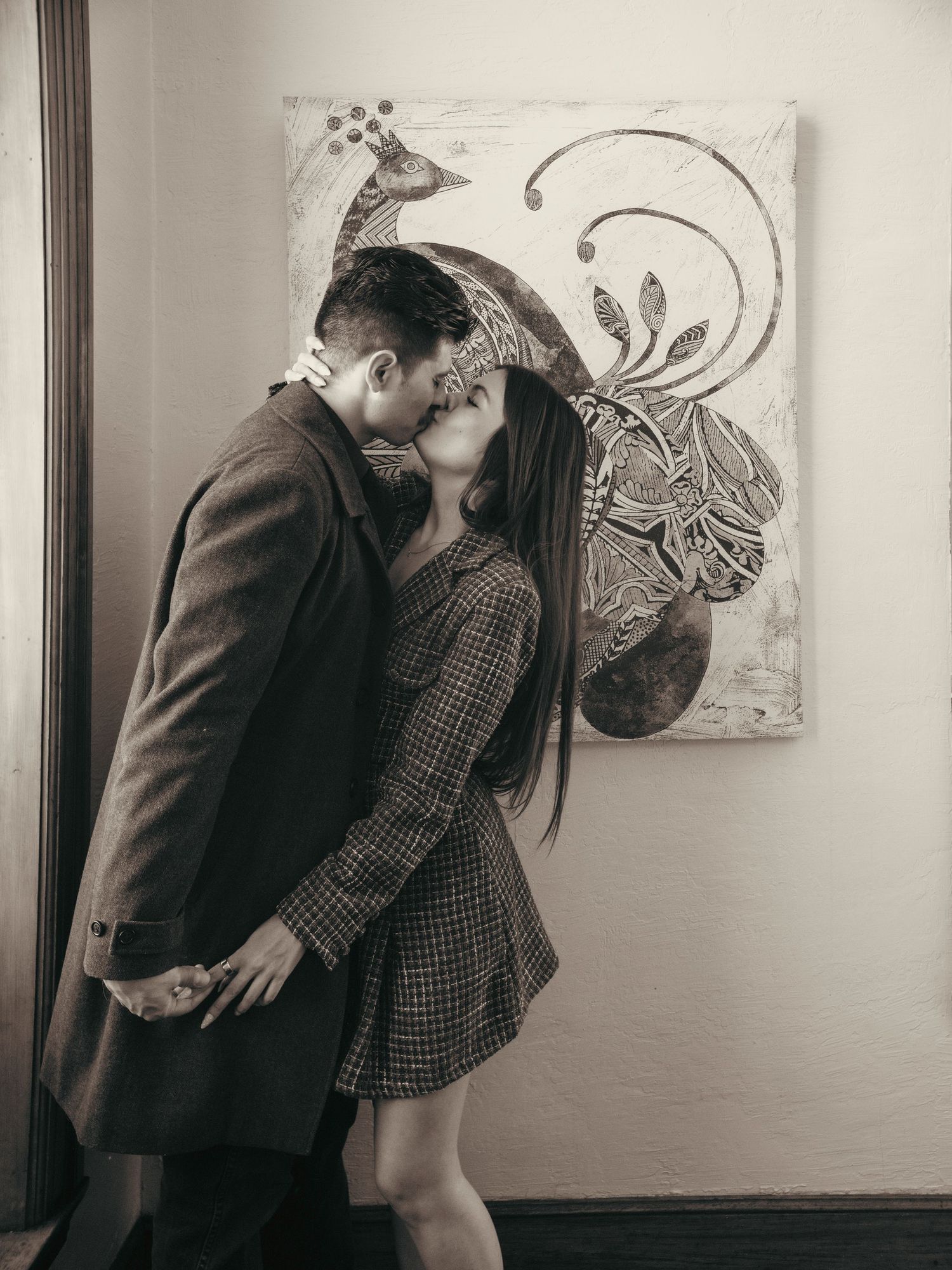 Couple sharing a romantic kiss in black and white photo with decorative peacock artwork on wall behind them.