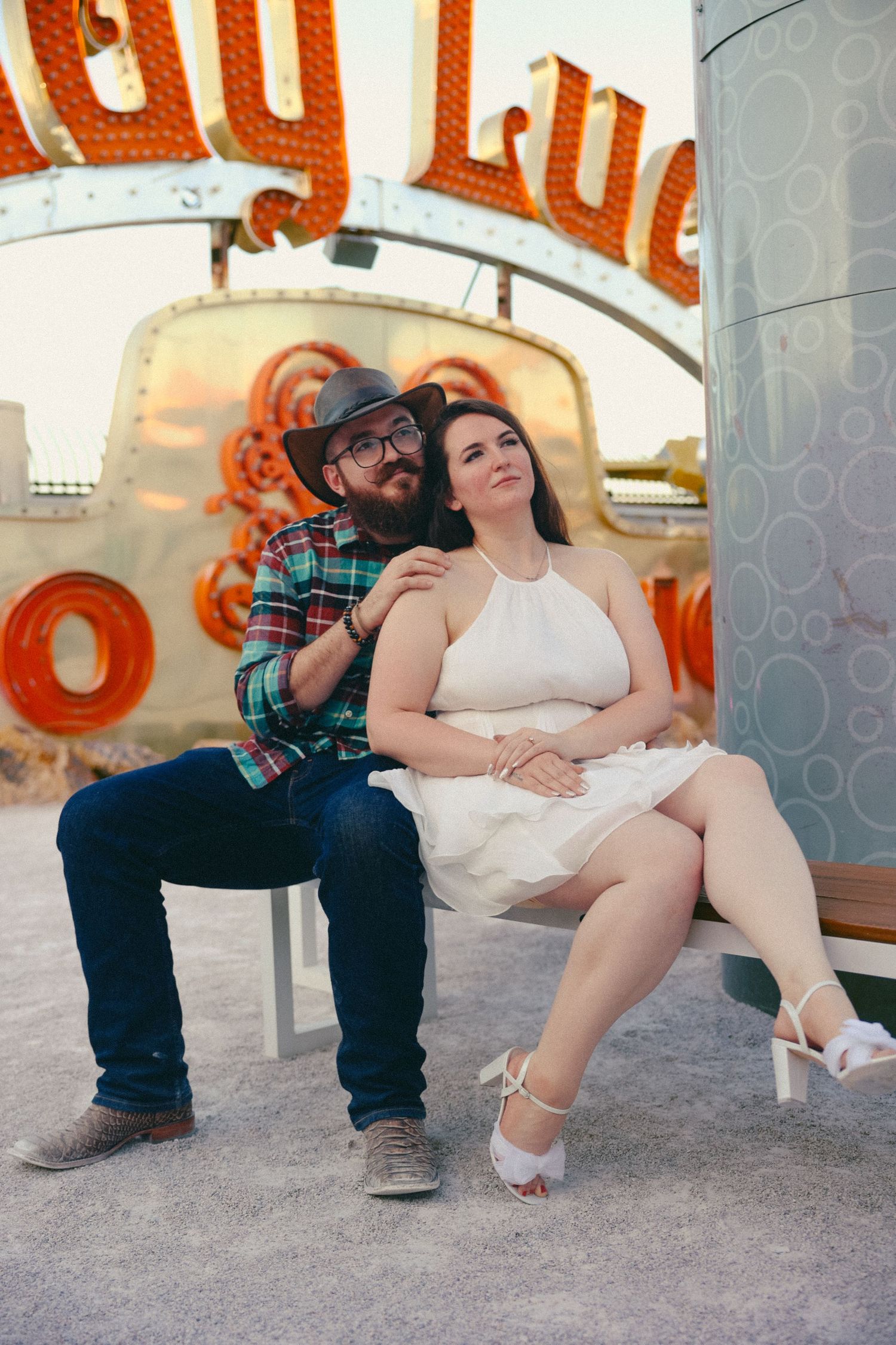 Couple sitting together at the Neon Museum in Las Vegas, man in plaid shirt and hat, woman in white dress.