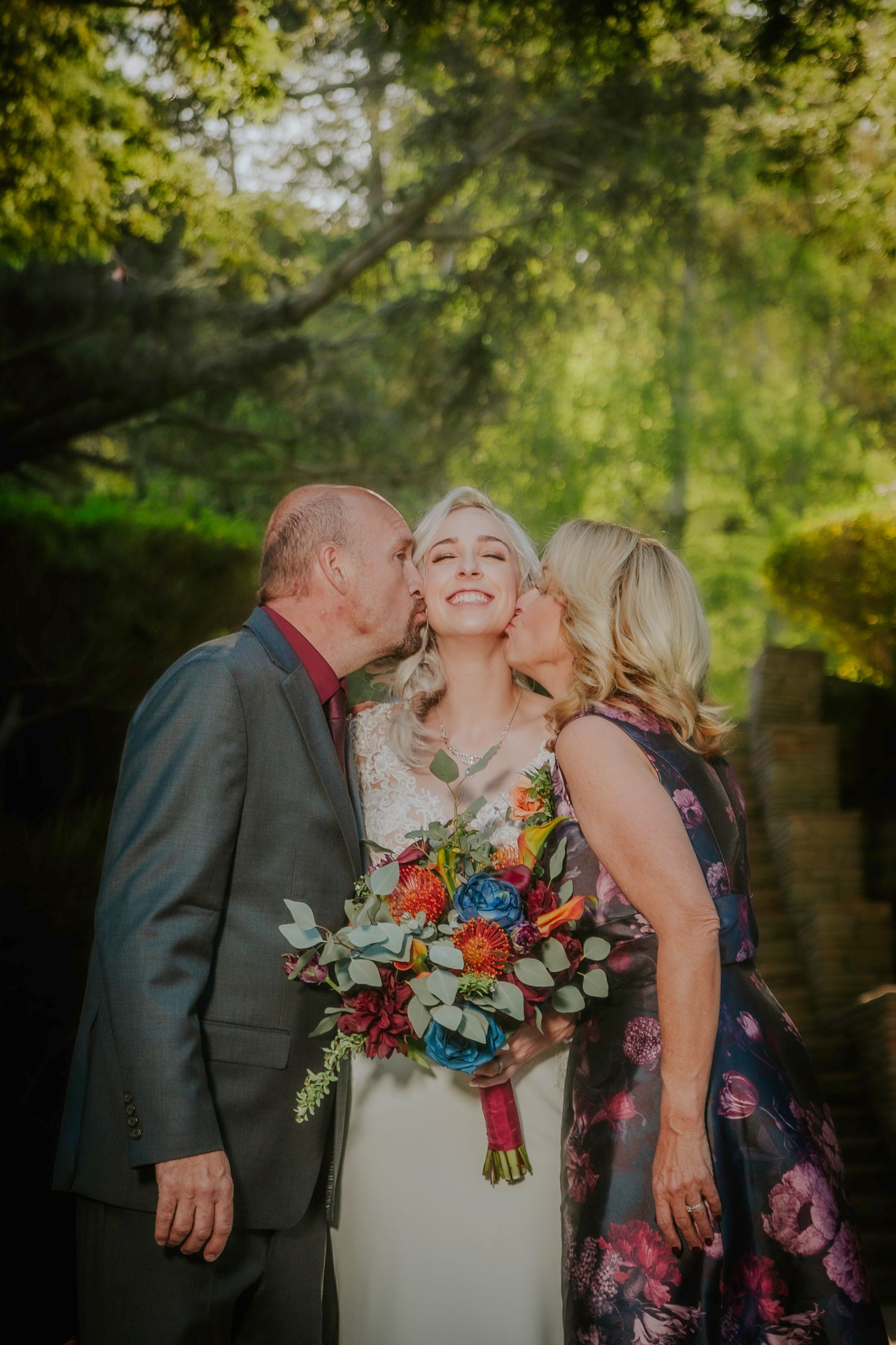 Bride smiling joyfully while parents kiss her cheeks, holding colorful bouquet outdoors at wedding.