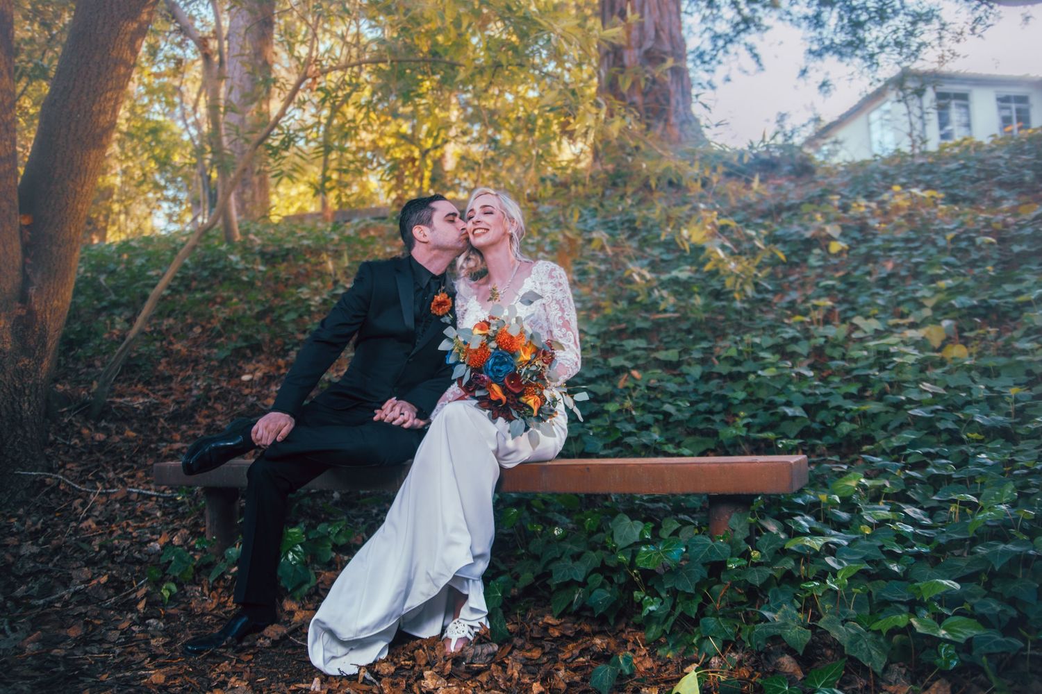 A bride and groom share a kiss on a wooden bench surrounded by autumn foliage in a romantic outdoor wedding setting.