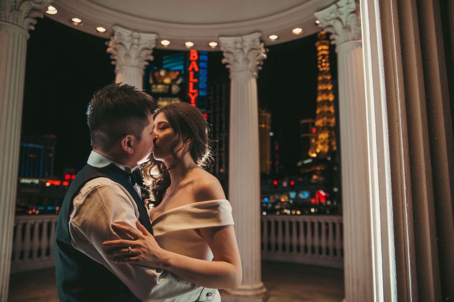 Bride and groom share a romantic kiss on a balcony overlooking the Las Vegas Strip at night.