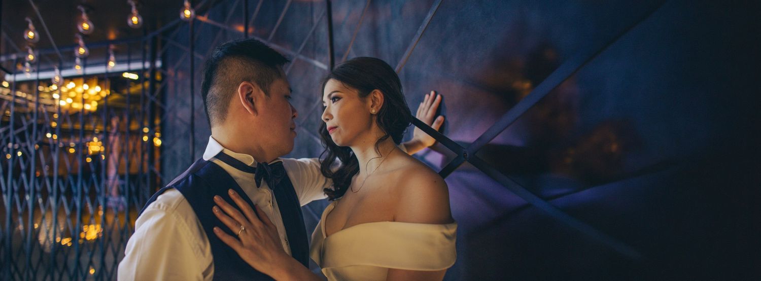 A couple shares a romantic moment in a dimly lit venue with warm bokeh lights, the woman in an off-shoulder gown.