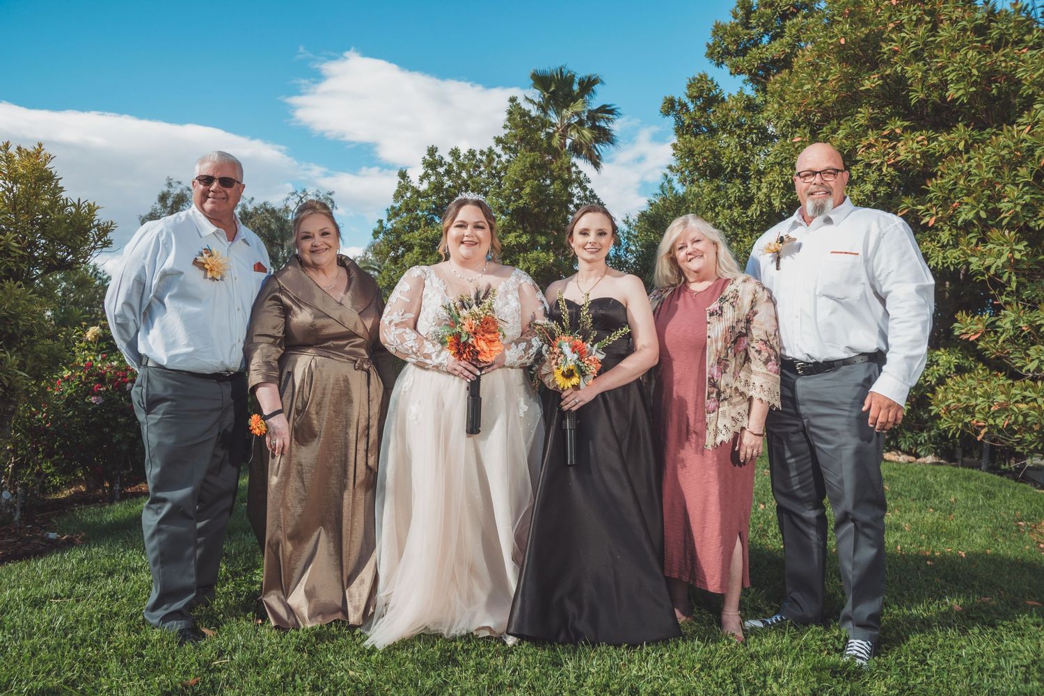 Wedding party of six poses outdoors on green lawn with trees and blue sky behind them.