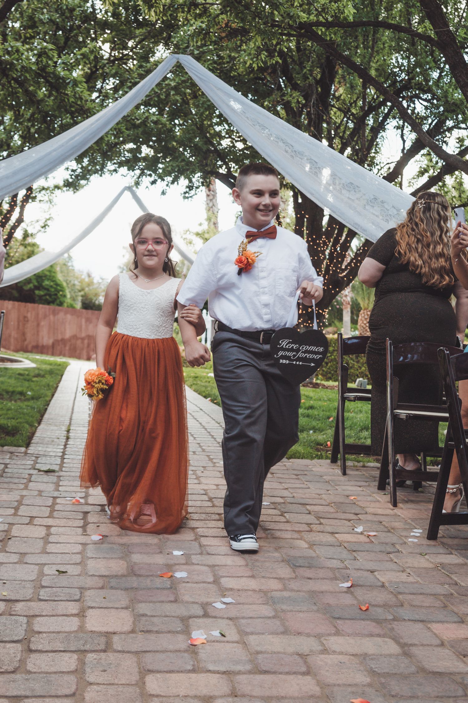Young flower girl in rust orange dress and ring bearer walk down outdoor wedding ceremony aisle.
