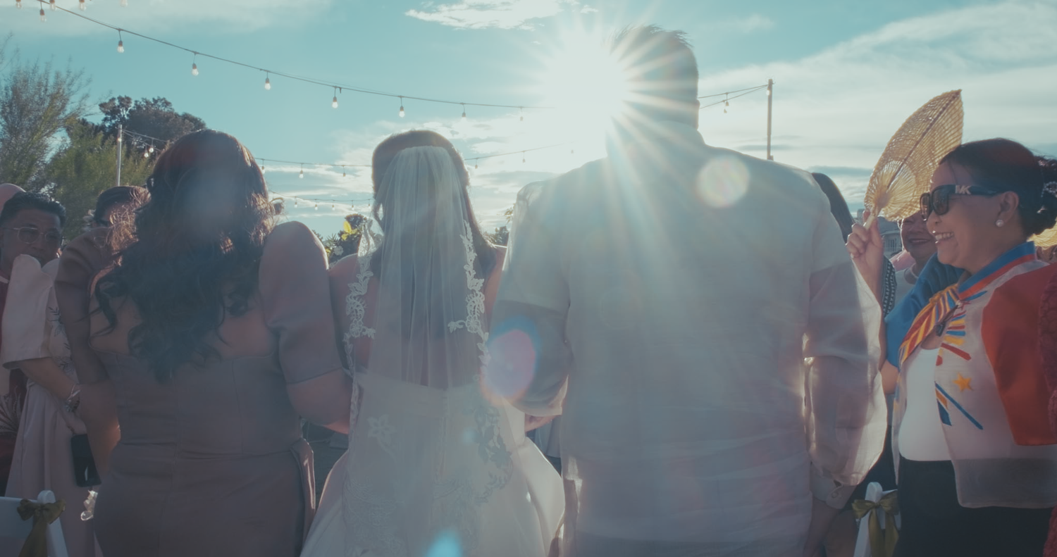 Bride in white gown walks through sunny outdoor wedding reception with guests gathered around.