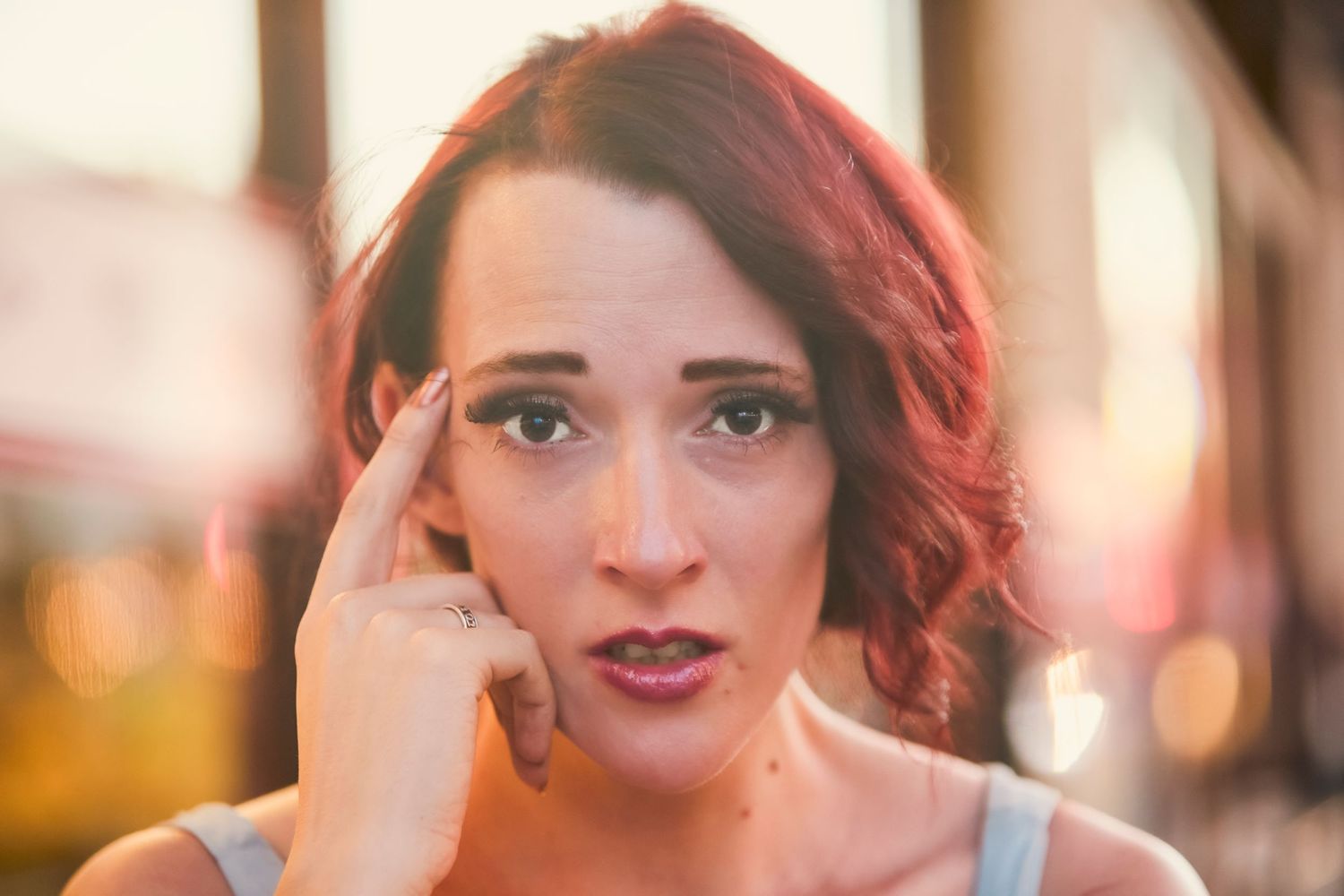 Young woman with red hair pointing to her temple with a surprised expression outdoors.