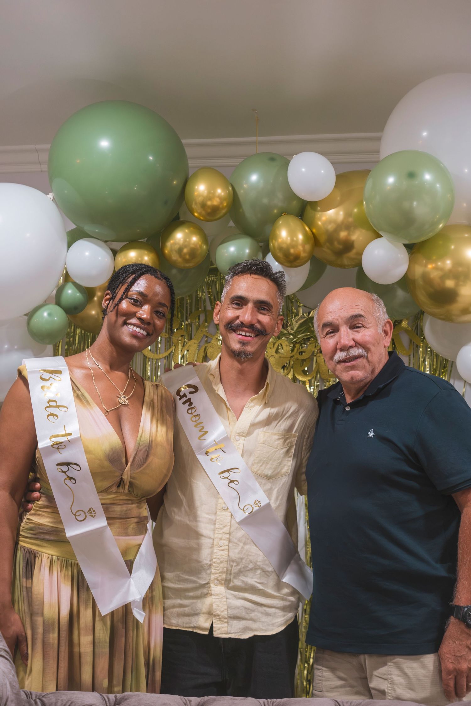 Couple wearing bride and groom sashes pose with older man at baby shower with green, gold, and white balloons.