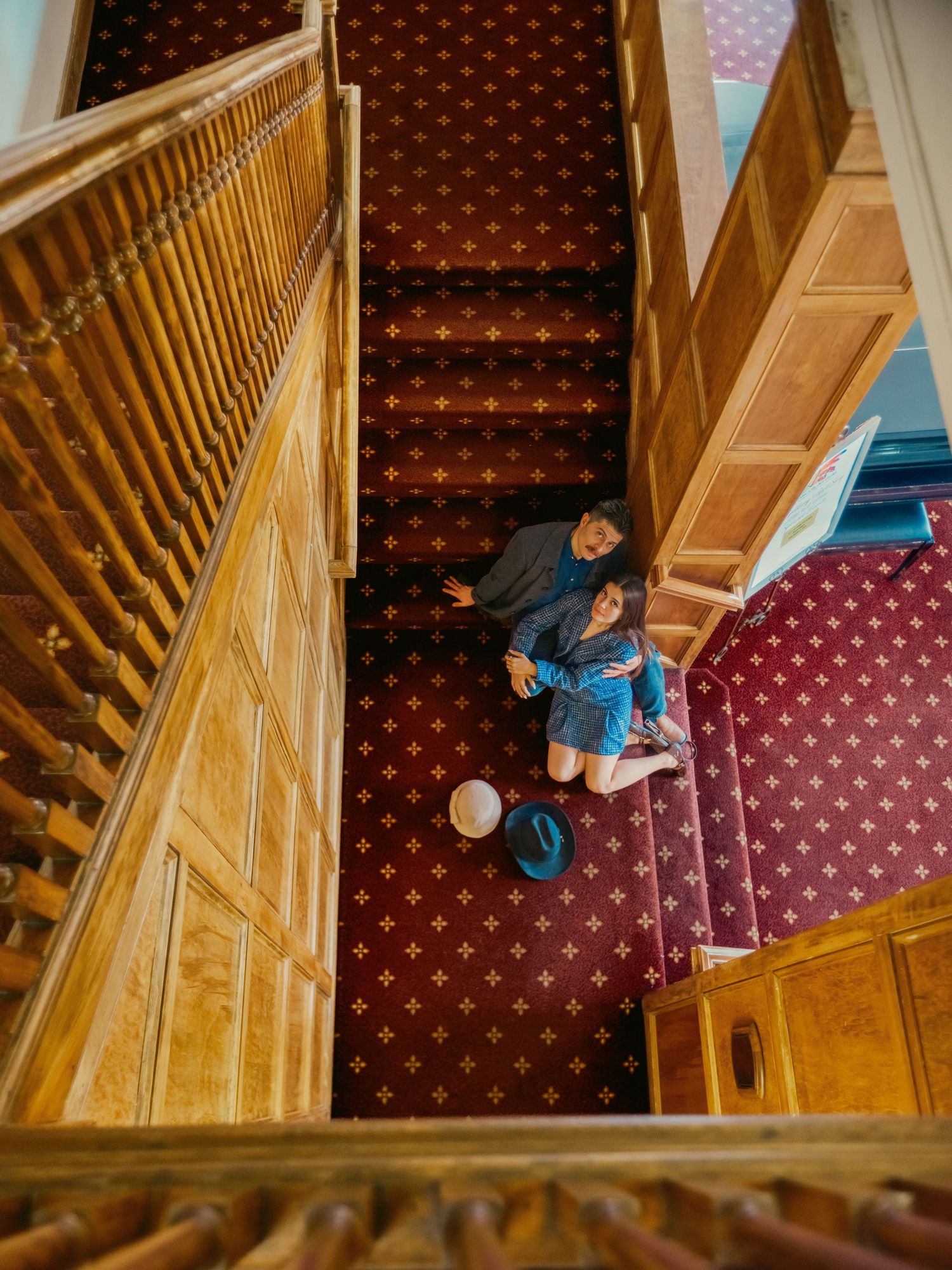 Aerial view of couple standing at base of grand wooden staircase with ornate red patterned carpet.