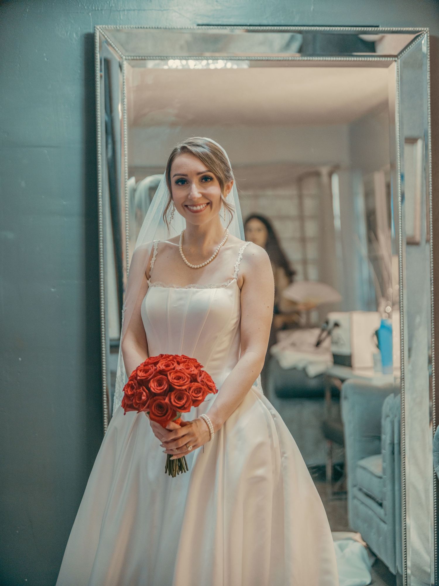 Smiling bride in white gown and veil holds red rose bouquet, standing in doorway with pearl necklace.