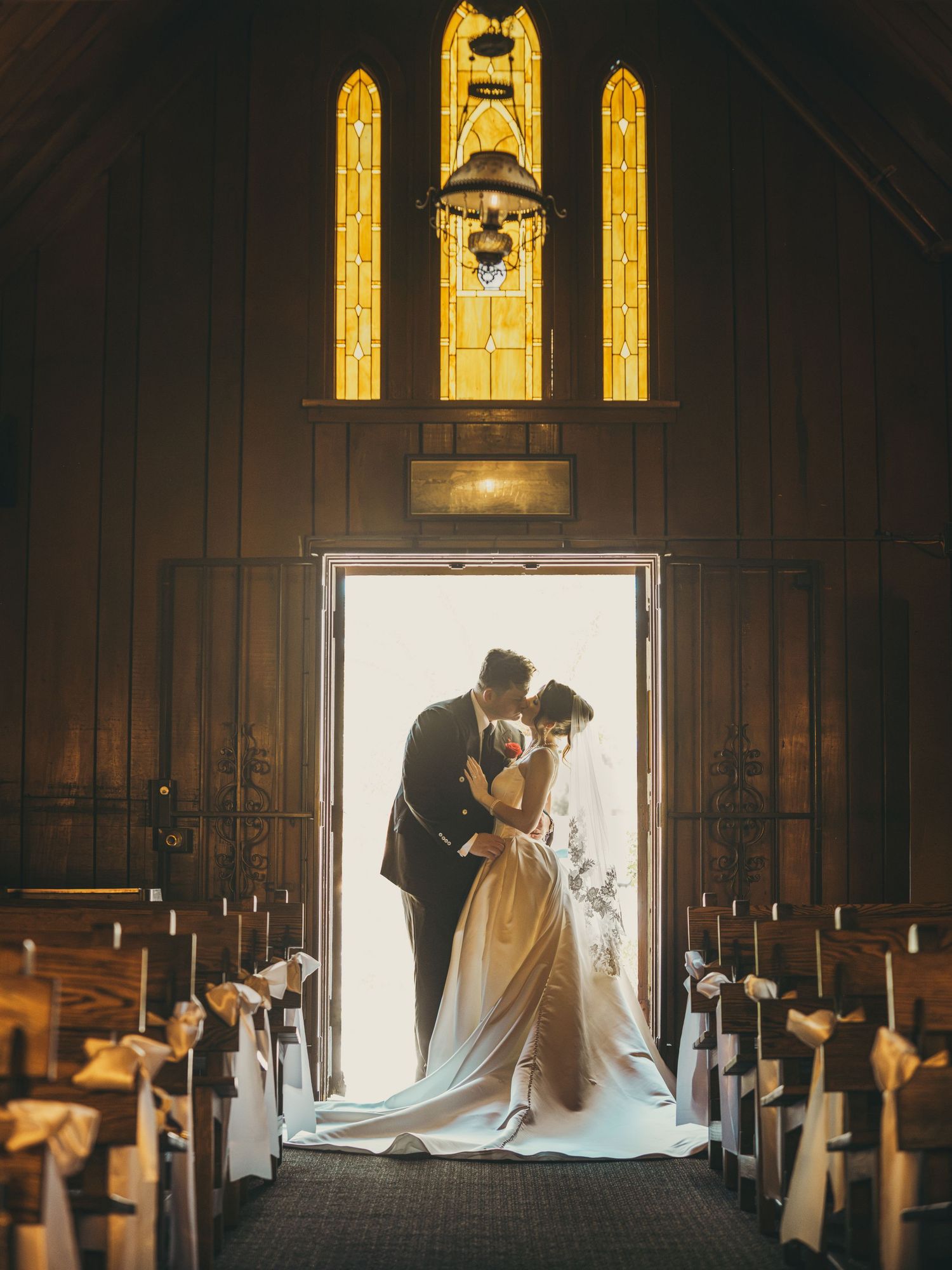 Bride and groom sharing a romantic kiss at church doorway with stained glass windows and candlelit pews.