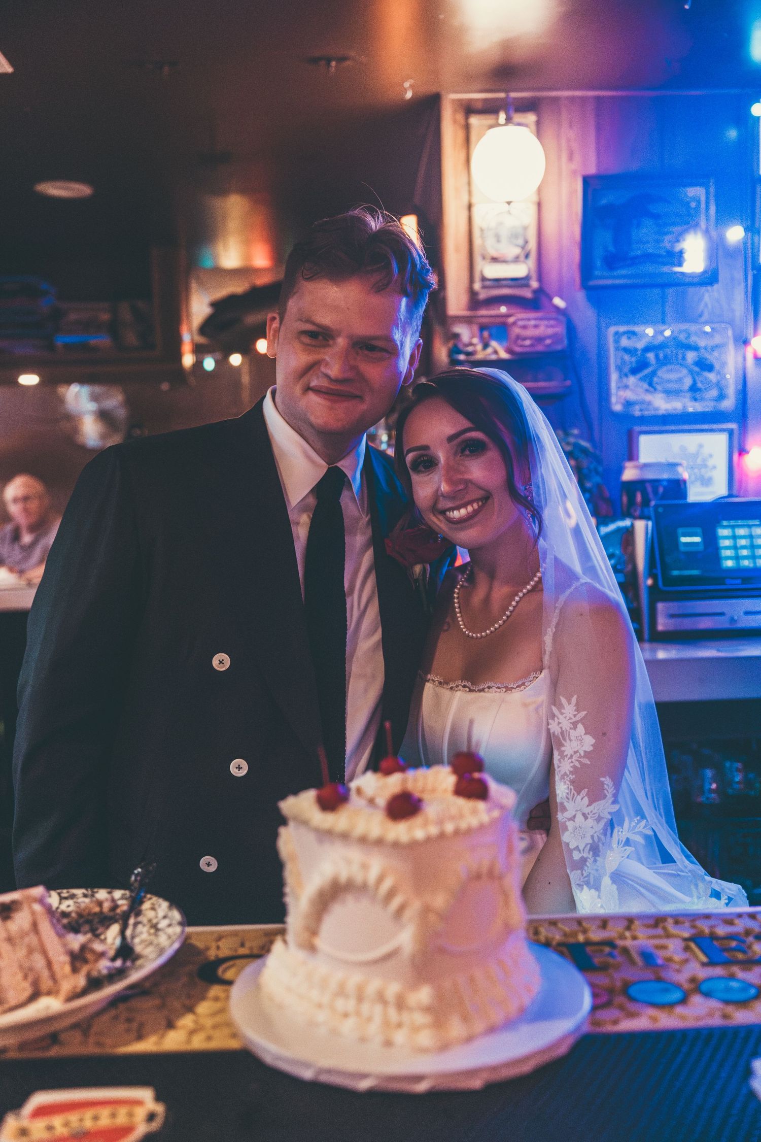 A smiling bride and groom pose together beside a white wedding cake in a warmly lit bar venue.