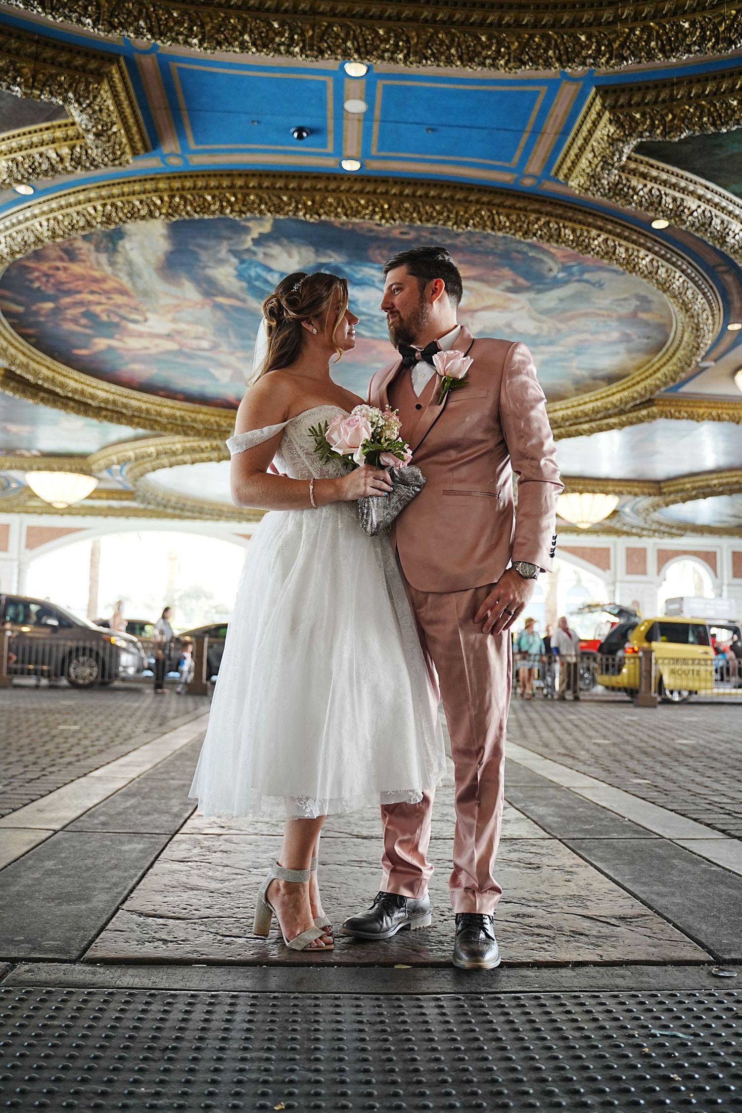 Bride in white tea-length dress and groom in pink suit share a romantic moment under ornate gold ceiling.