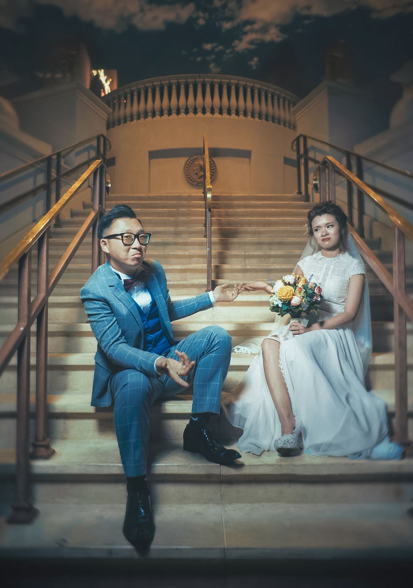 Bride and groom sitting on grand staircase, groom in blue suit holding hands with bride in white gown holding bouquet.