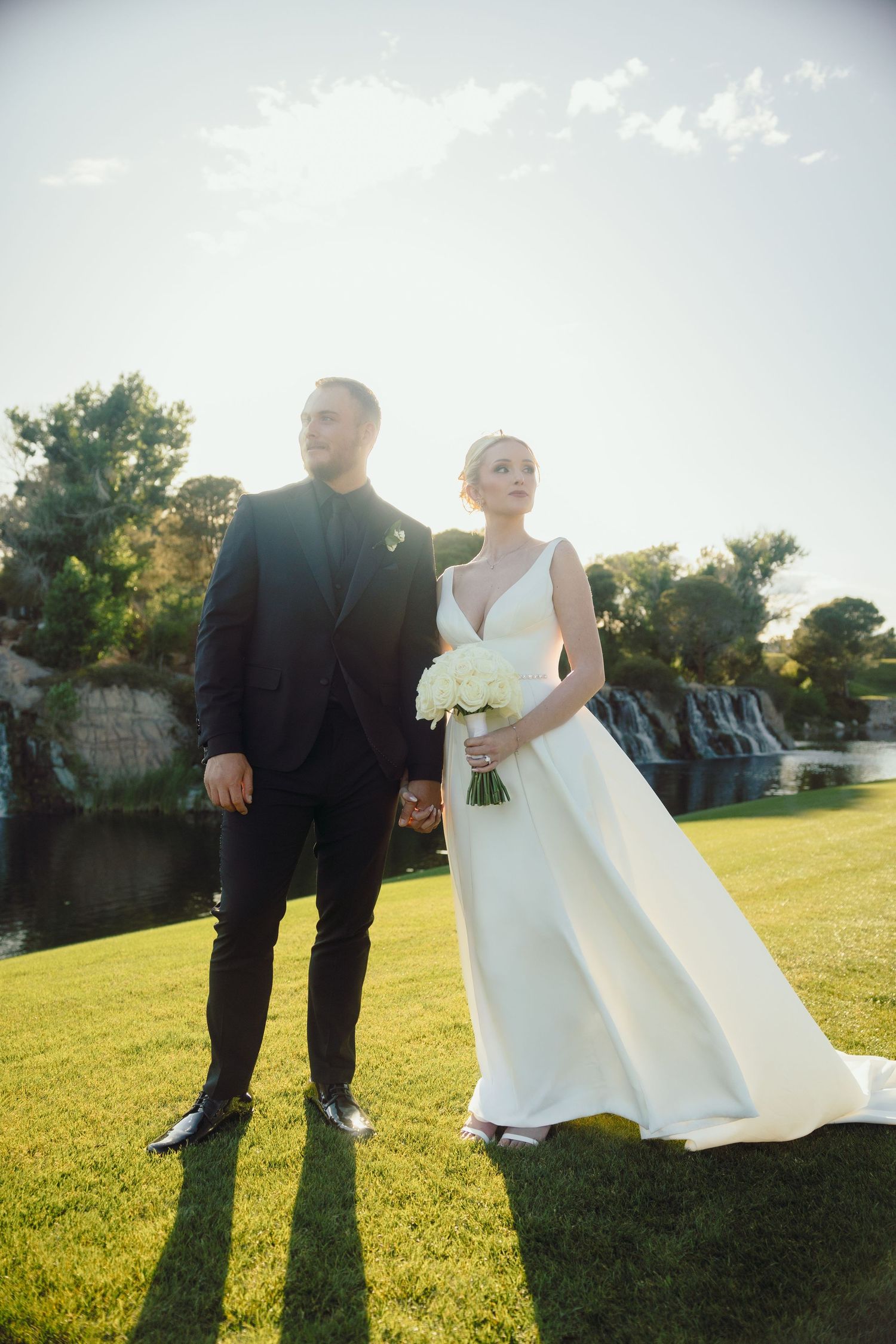 Bride holding bouquet and groom standing on lawn with waterfall in background.