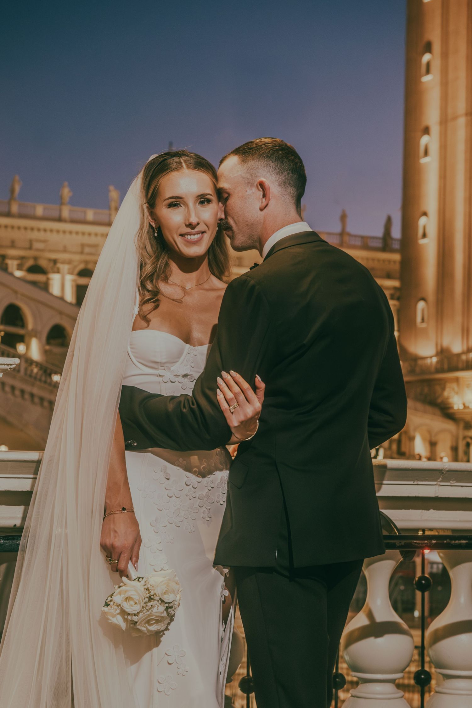 Bride smiles warmly as groom embraces her on a balcony with the illuminated Venetian resort behind them.