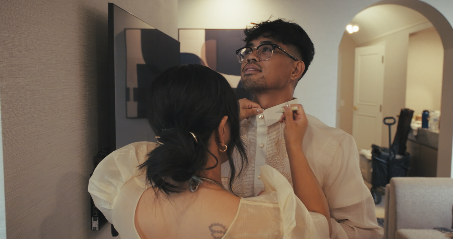 A woman adjusts a groom's collar in a mirror reflection during wedding day preparations.