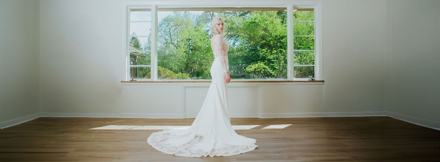A bride in a white mermaid wedding gown stands elegantly by large windows in a bright, wood-floored room.
