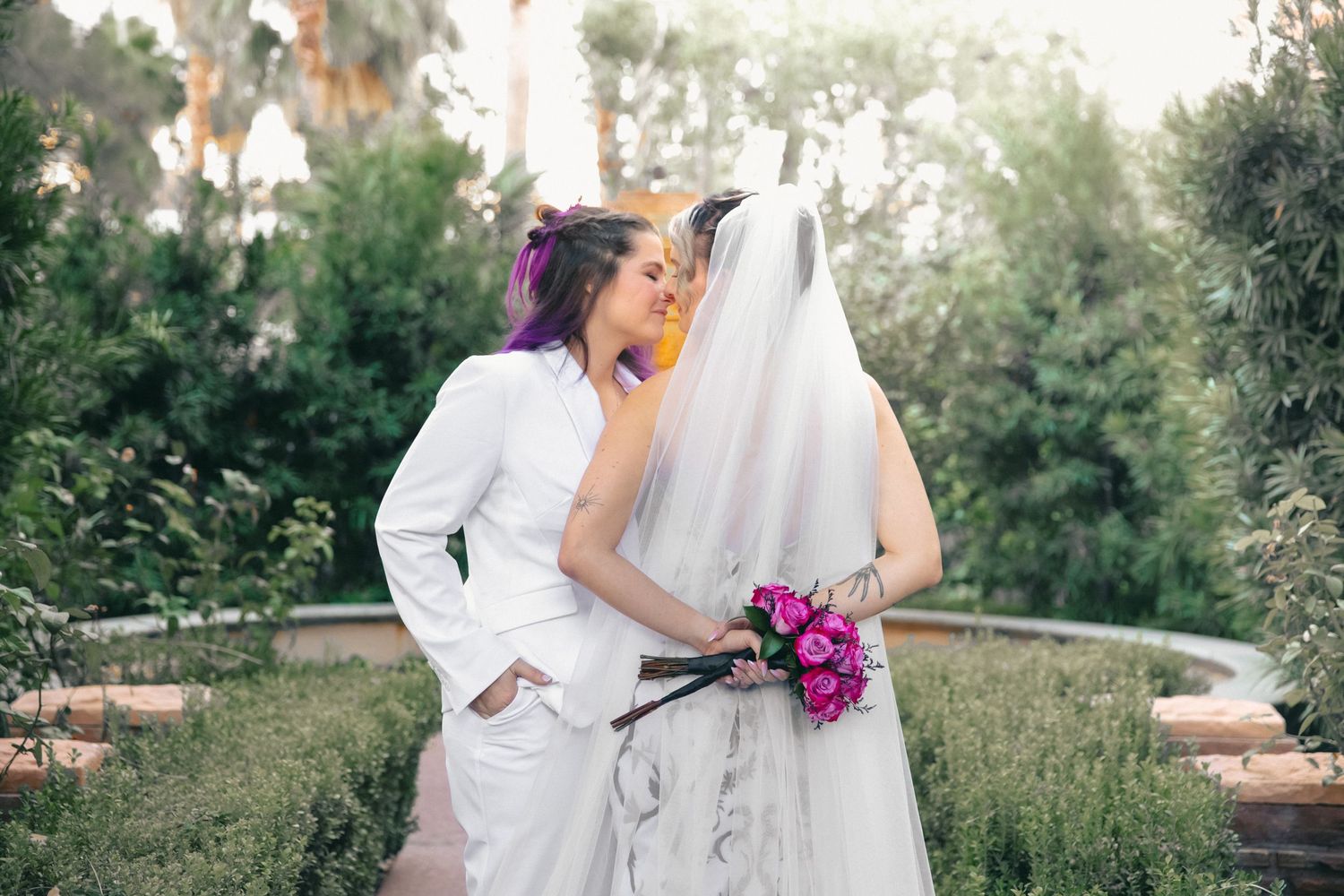 Two brides share a kiss outdoors, one in white suit with purple hair holding pink bouquet.