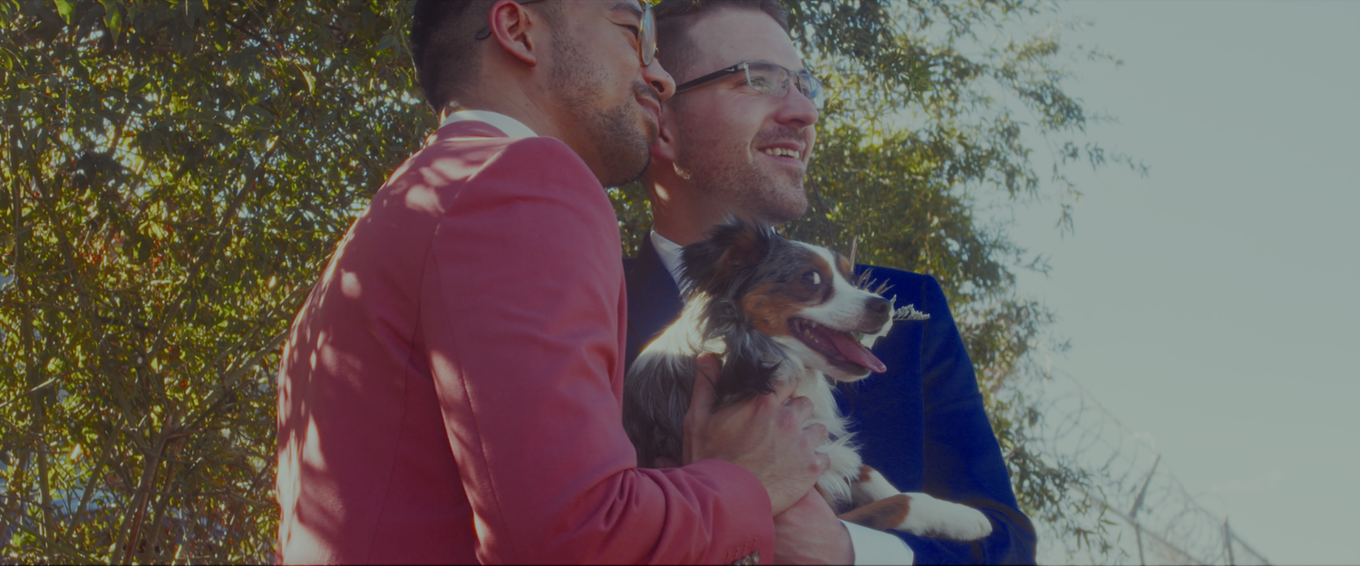 Two smiling men holding an Australian Shepherd dog outdoors on a sunny day.