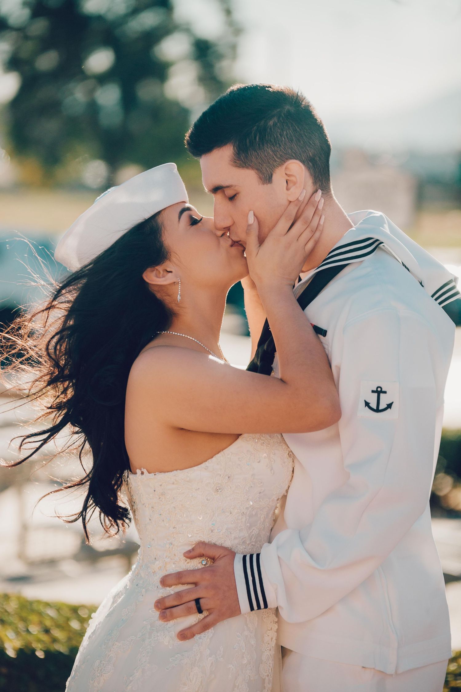 A bride and groom in a Navy sailor uniform share a romantic kiss outdoors on their wedding day.