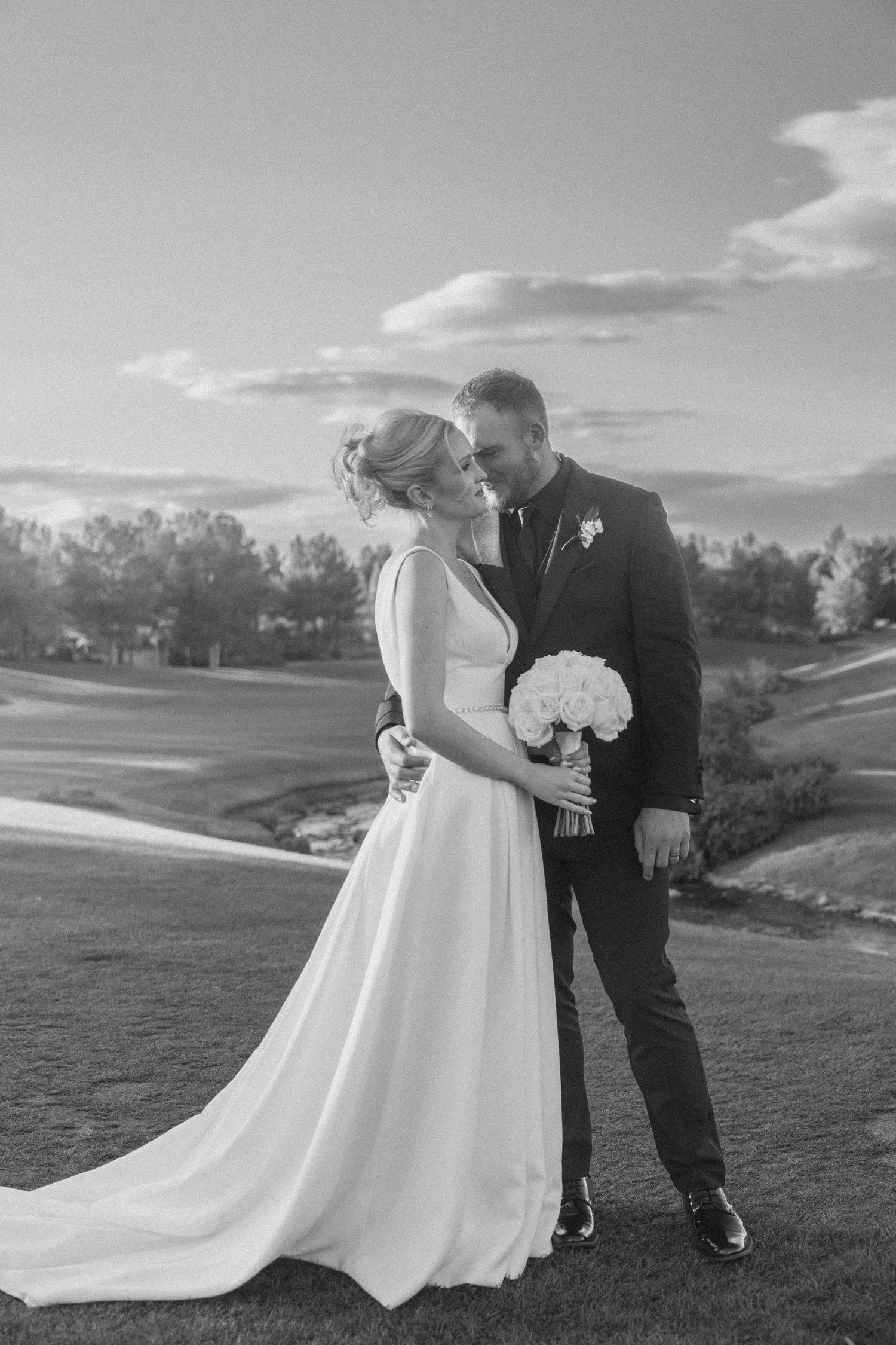 Black and white photo of bride and groom embracing on golf course with bouquet.