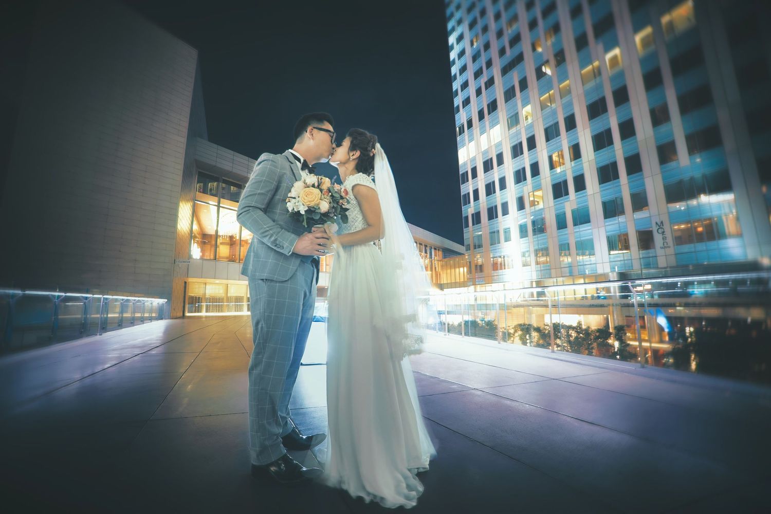 Bride and groom share a romantic kiss on a rooftop at night with illuminated city buildings in the background.