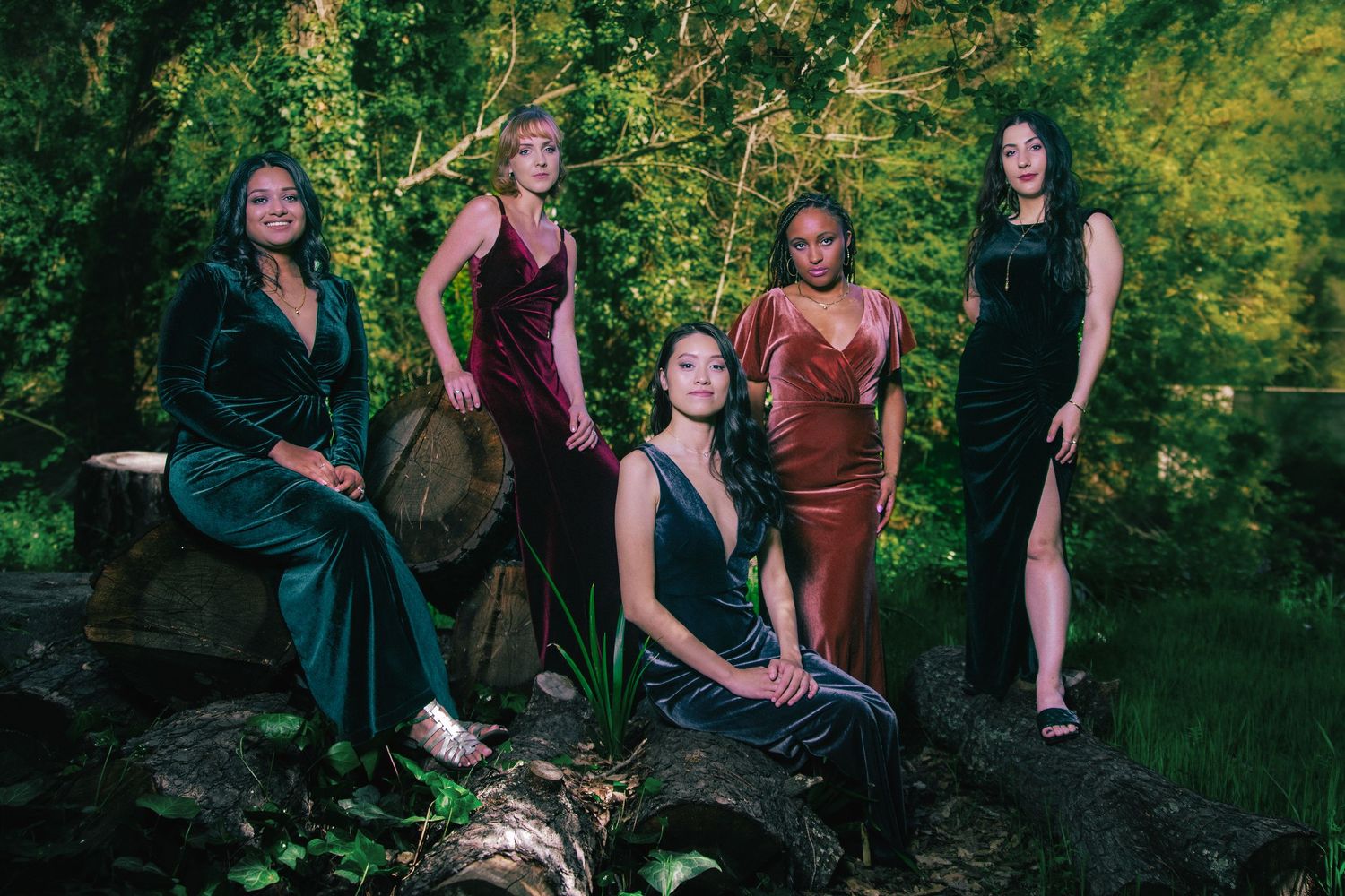 Five women in elegant velvet gowns pose gracefully among lush green trees and logs in an outdoor forest setting.
