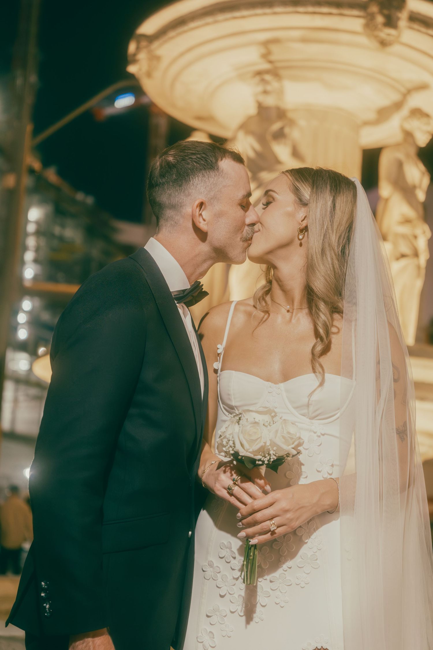Newlyweds share a kiss in front of glowing fountain at night, bride holding white rose bouquet in Vegas.