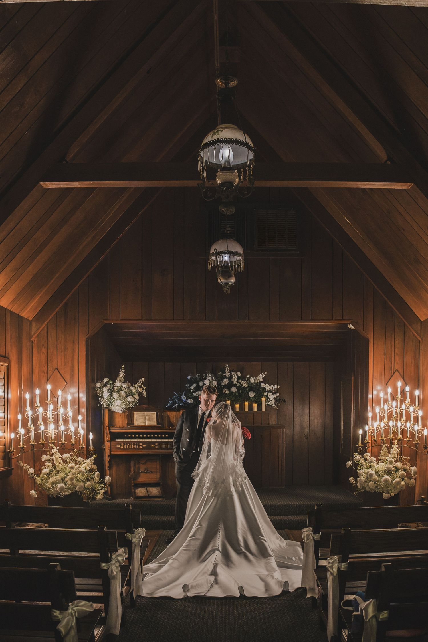 A bride and groom stand at the altar of a candlelit wooden chapel adorned with white floral arrangements.