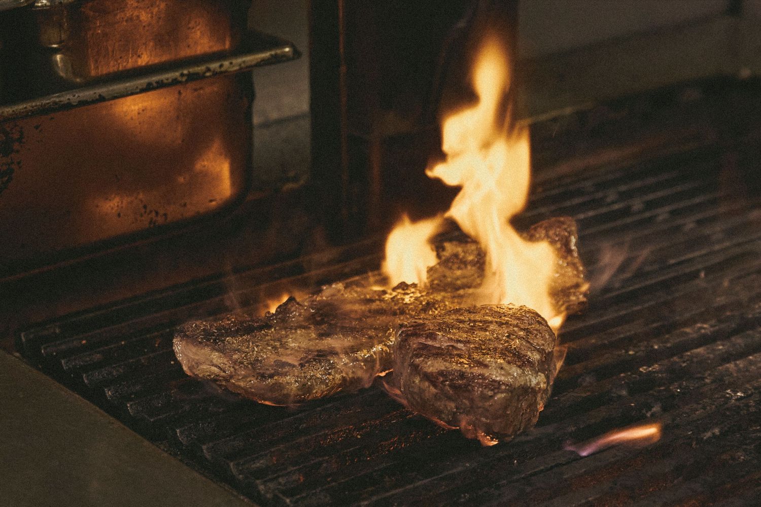 Two juicy steaks grilling over open flames on a metal grill grate, with fire rising dramatically around the meat.
