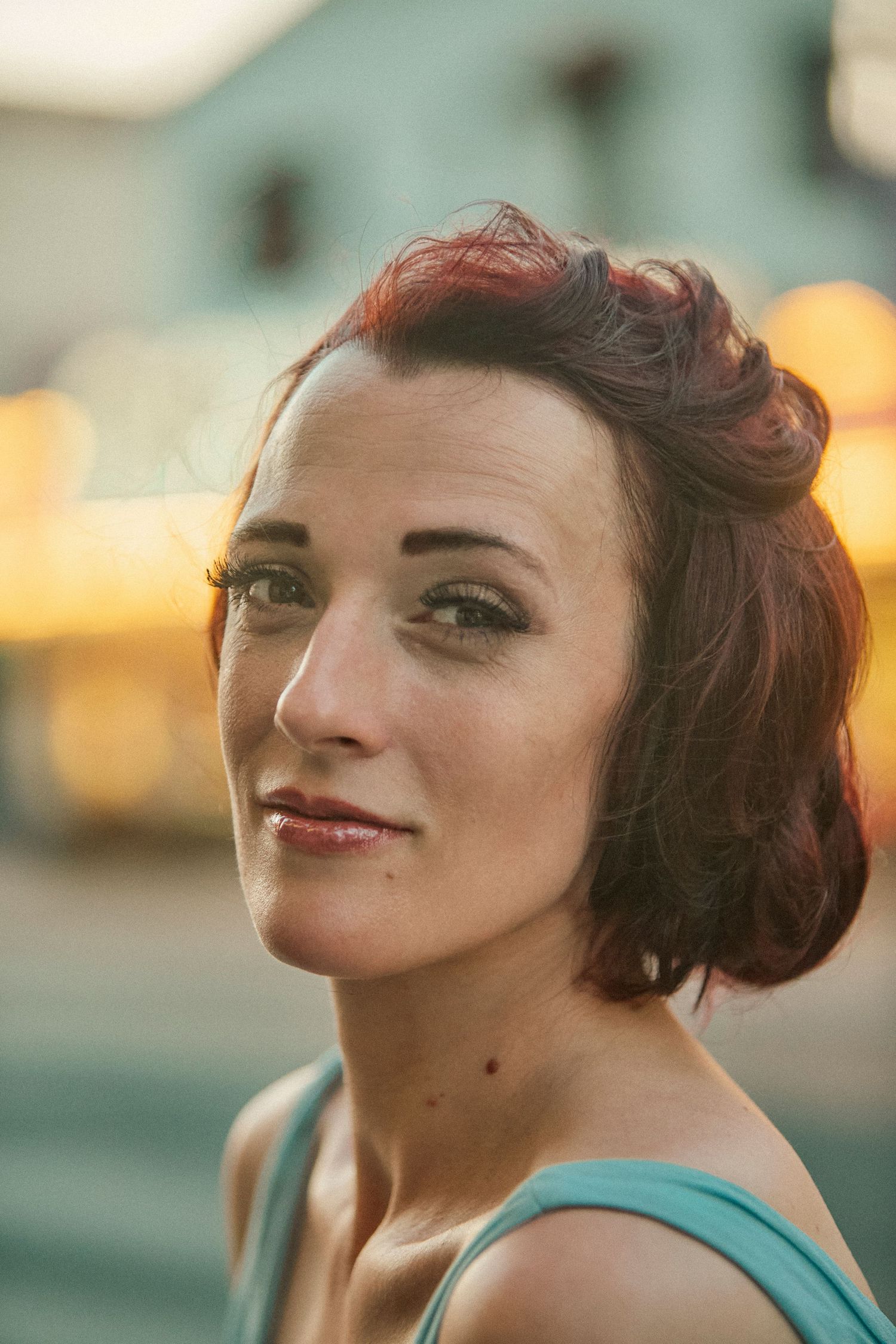 A young woman with red upswept hair and teal top poses outdoors at golden hour with bokeh lights in background.