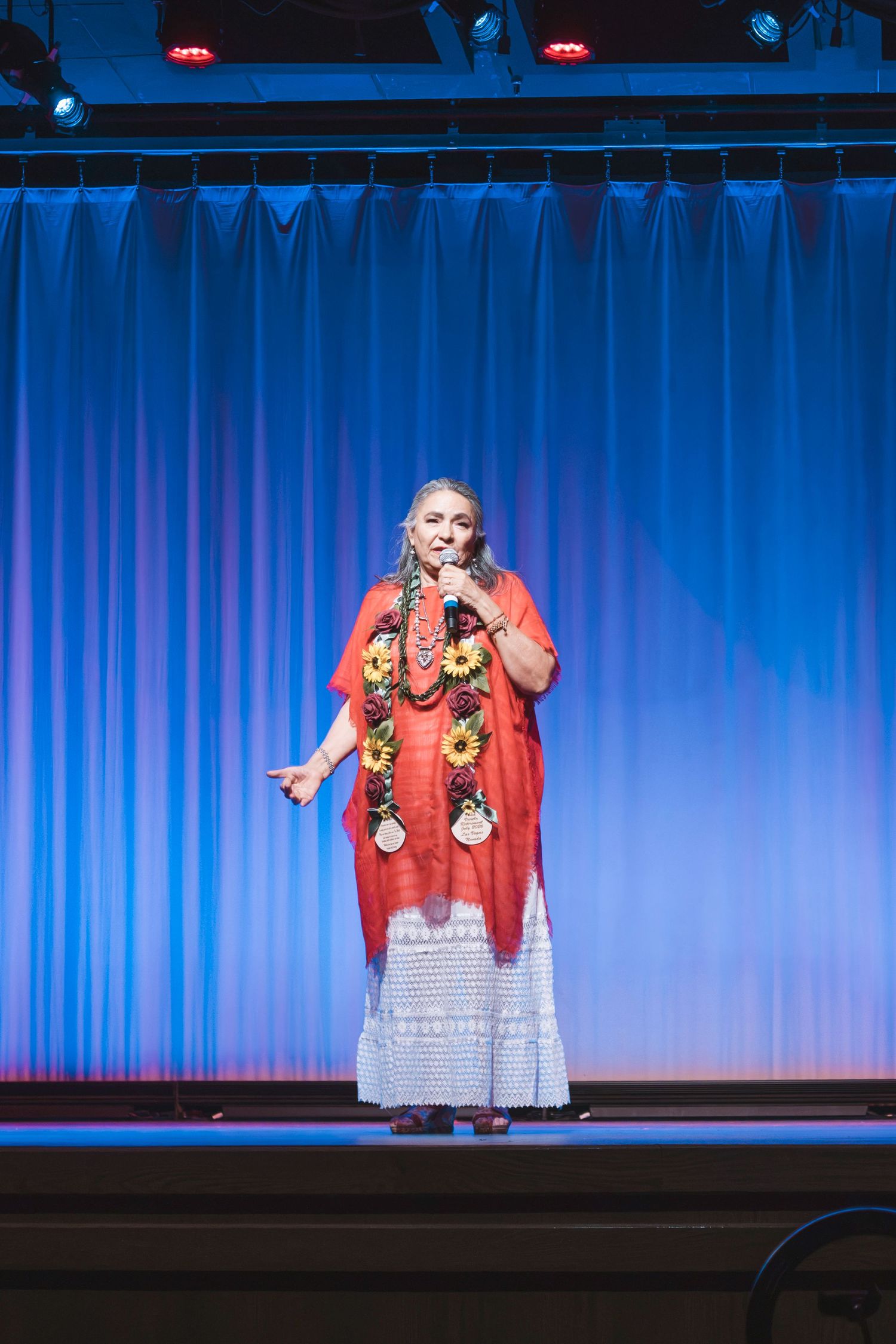 Woman in red kimono with floral lei performs on stage with microphone before blue curtain backdrop.