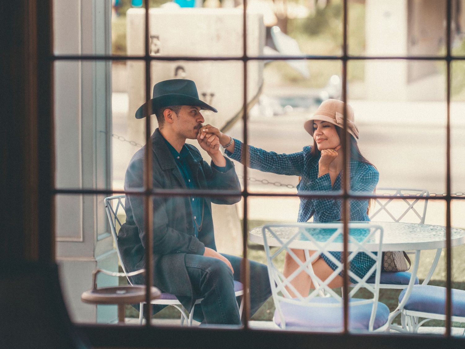 Couple sitting at outdoor cafe table, man in cowboy hat and woman in sun hat enjoying a romantic moment.