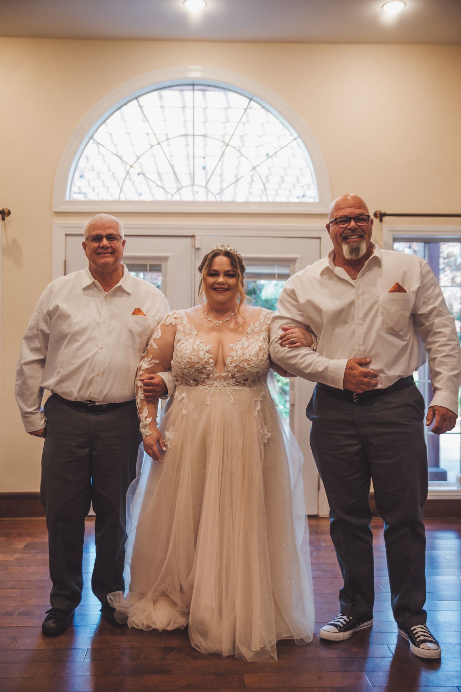 Bride in lace wedding dress stands between two men in white shirts before arched window.