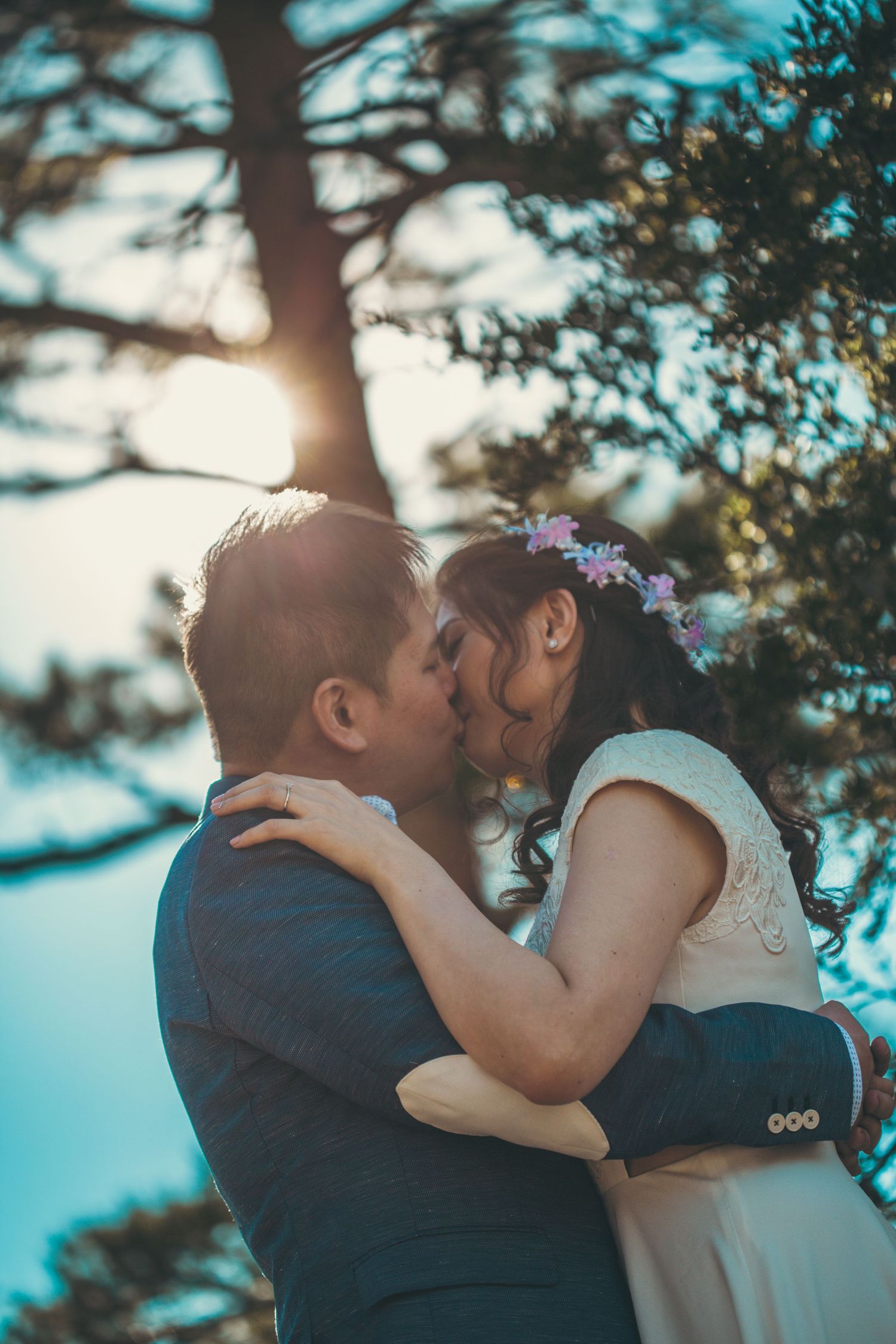 Bride and groom sharing a romantic kiss under a sunlit tree, bride wearing floral hair accessories and white dress.