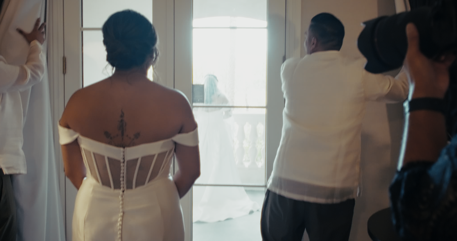 A bride in a white corset gown stands before bright doors surrounded by guests.