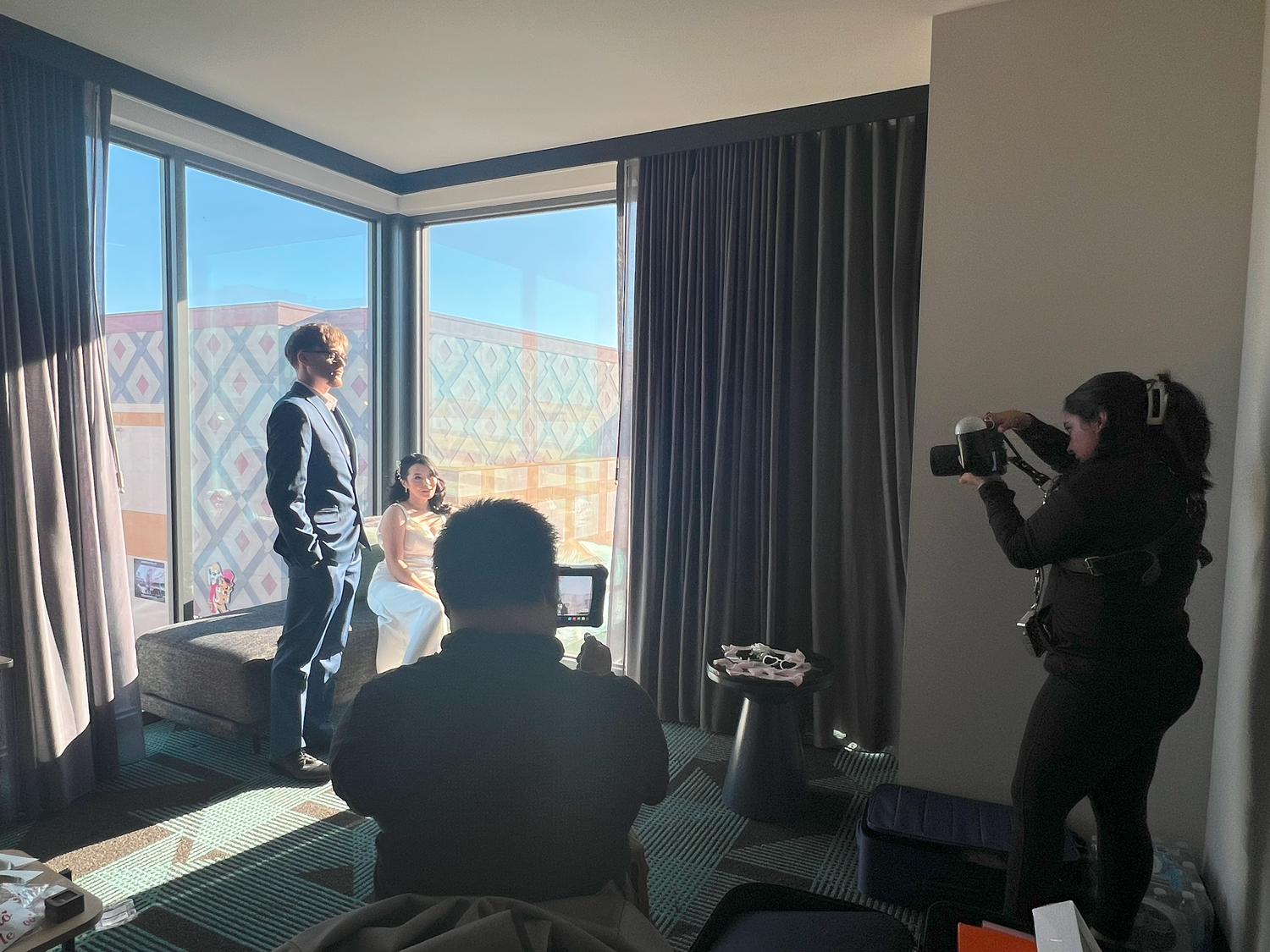 Photographer capturing a couple posing by large corner windows in a modern hotel room with city views.